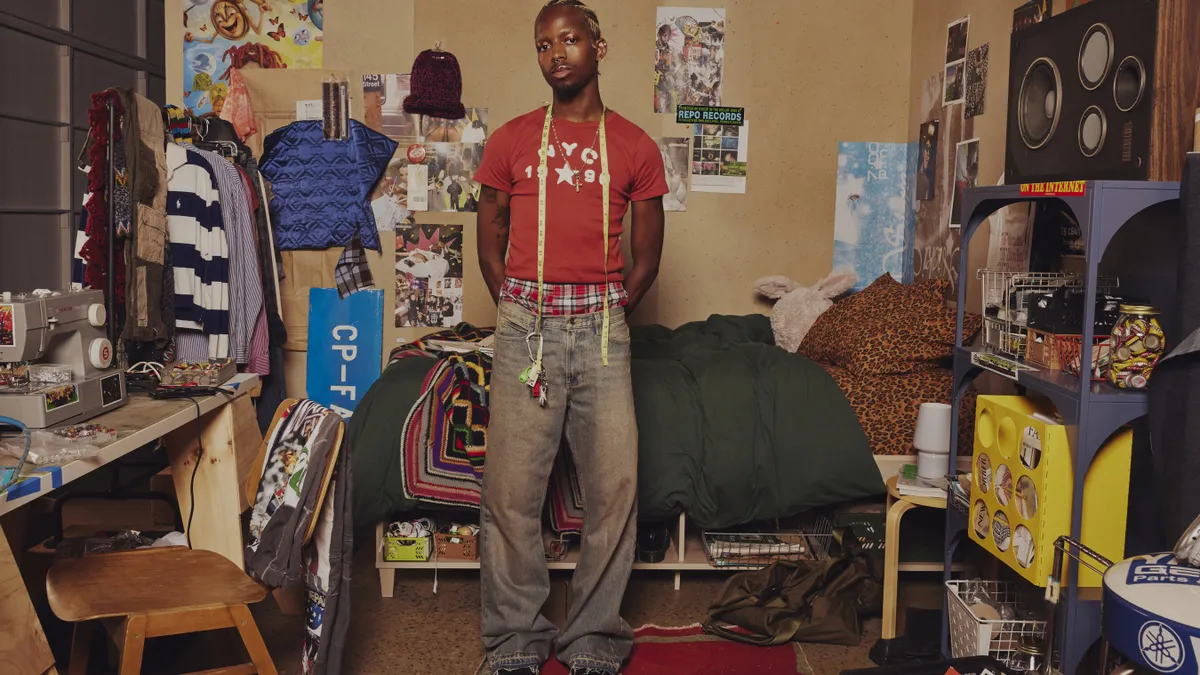 A man stands in a room reminiscent of a college dorm wearing jeans and a red NYC t-shirt.