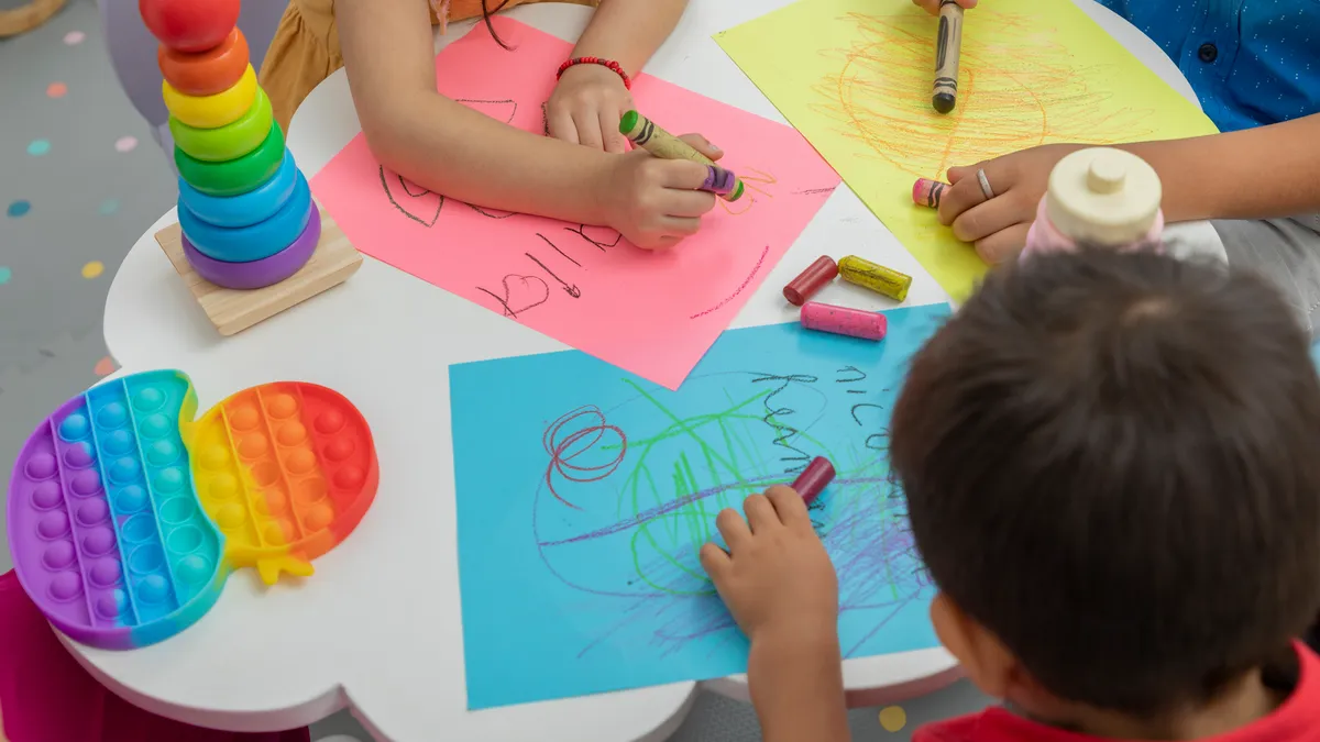 An overhead shot of a small table with three children using crayons to color on paper. A stacking toy and popper toy are also on the table.