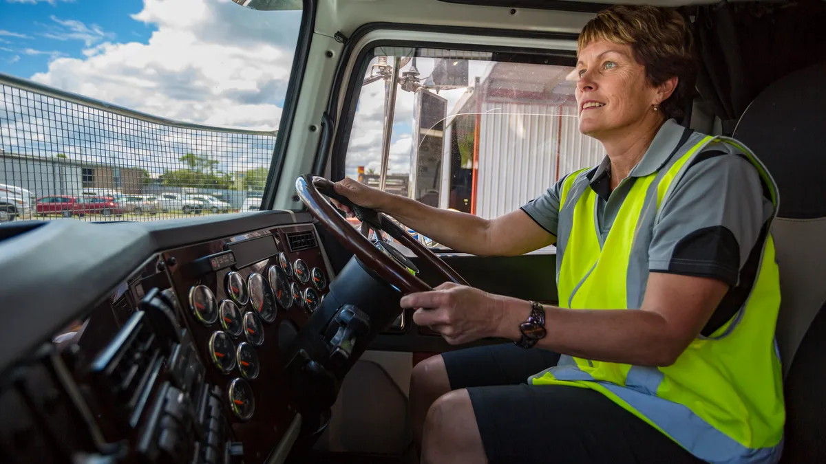 A female driver holds the wheel of a truck wearing high visibility clothing.