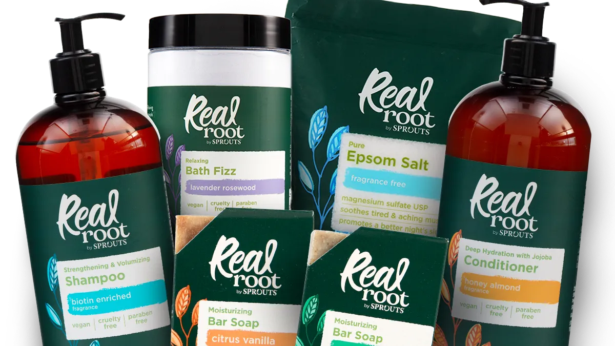 Array of Sprouts Real Root products, including bottles of shampoo and conditioner, bar soaps, bath fizz and Epsom salt all in dark green packaging against a dark gray background.