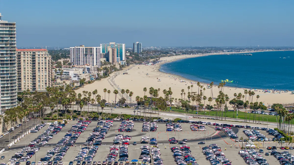 A parking lot full of cars and buildings line a sandy beach and blue water.