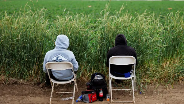 Two individuals sit on folding chairs outdoors, with lunch bags between them.