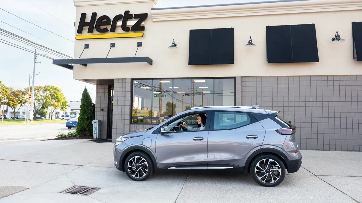 A gray four-door crossover vehicle with a woman seated with her hand on the steering wheel is parked in front of a Hertz building.