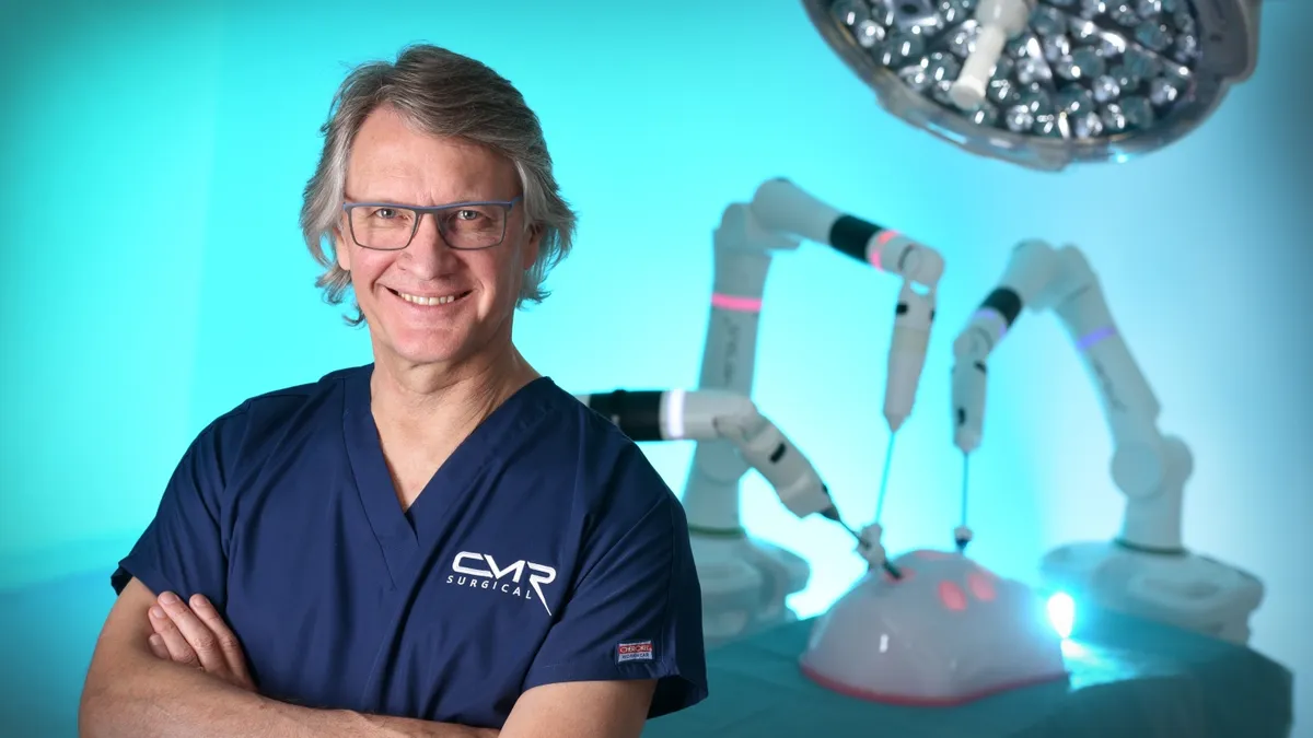 CMR Surgical Co-founder and CMO Mark Slack stands with his arms crossed in front of a Versius robot.