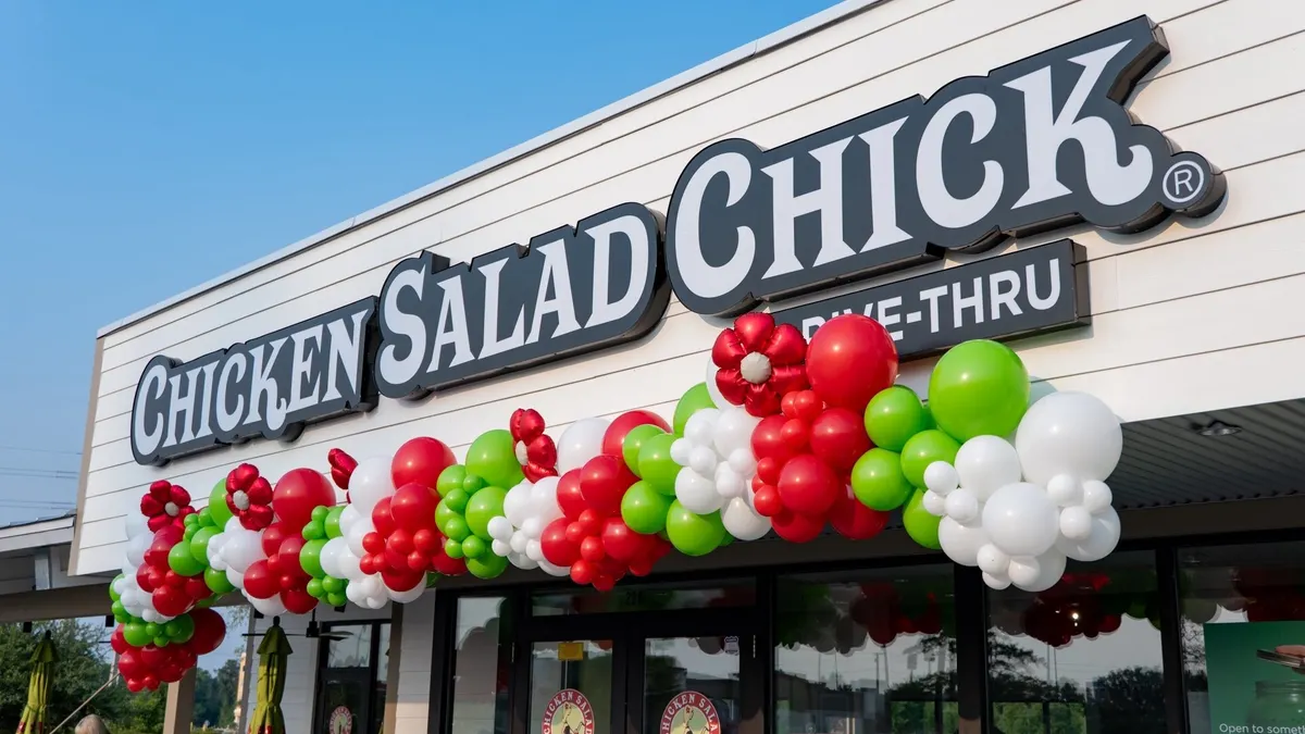 An photograph of a building sign that says Chicken Salad Chick drive-thru with red and green balloons underneath.