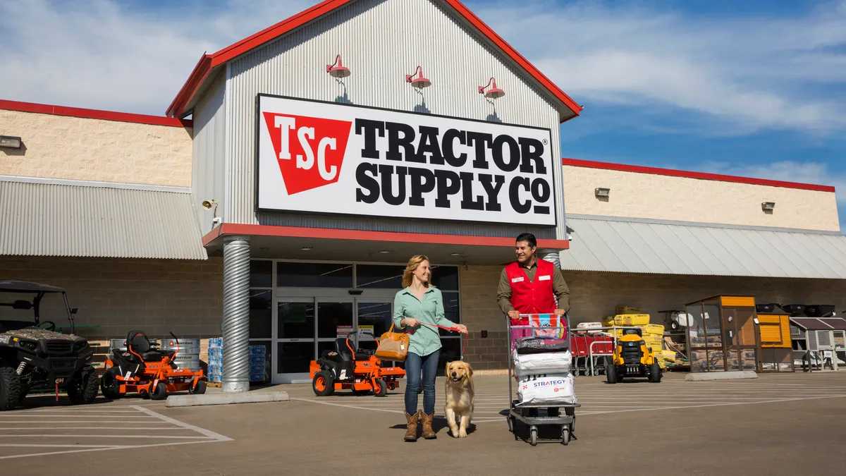 Two smiling people, one with a dog on a leash and the other with a shopping cart, walk away from a store entrance.