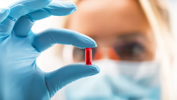 A scientist with protective eyeglasses and mask holding a red transparent pill with fingers in gloves in the pharmaceutical research laboratory