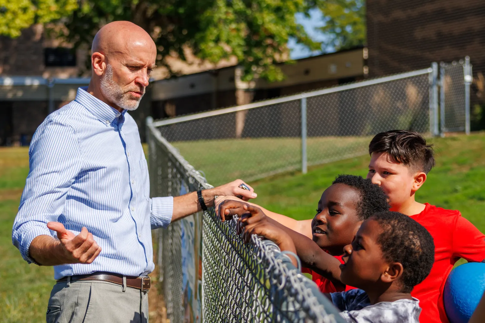 A man superintendent speaks with elementary students standing next to a fence outside of their school on a sunny day.