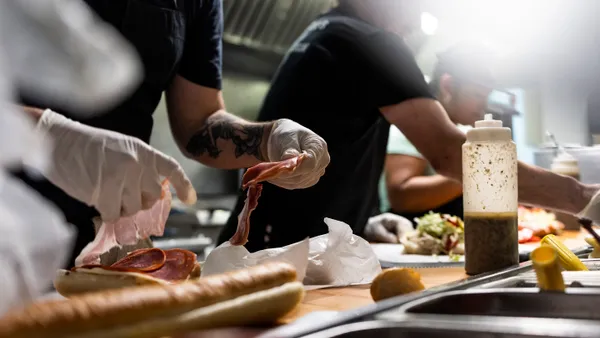 Sandwiches sit on a counter as workers as ingredients.