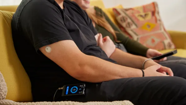A person sits on a couch with a white patch on their upper arm and a rectangular device with a screen on their hip.