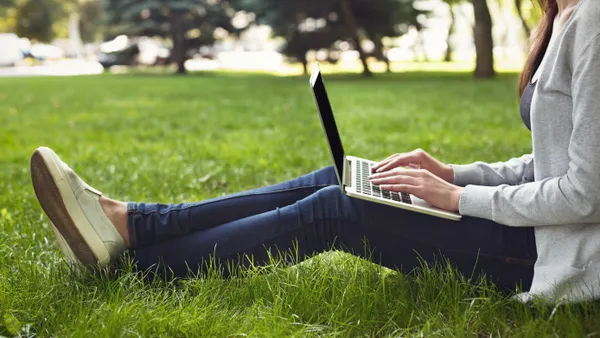 A person sits in the grass with a laptop on their lap and hands rested on the keyboard