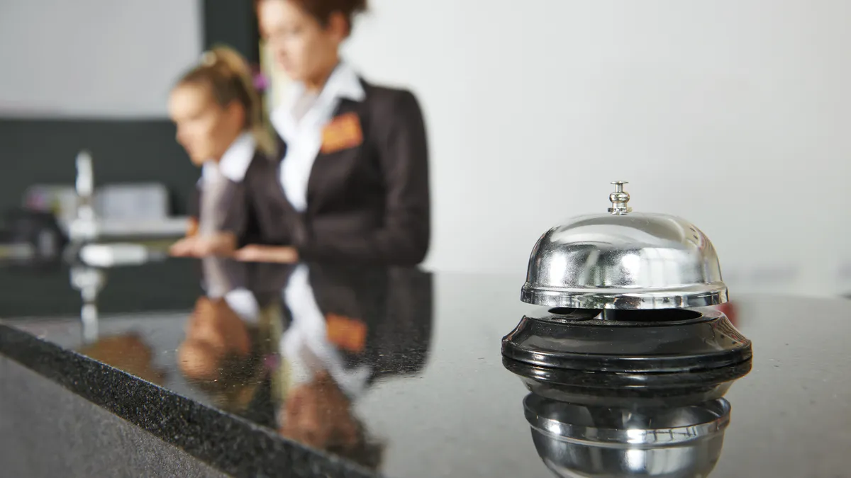 A close-up shot of a bell in a hotel lobby, with two workers pictured out of focus in the background.