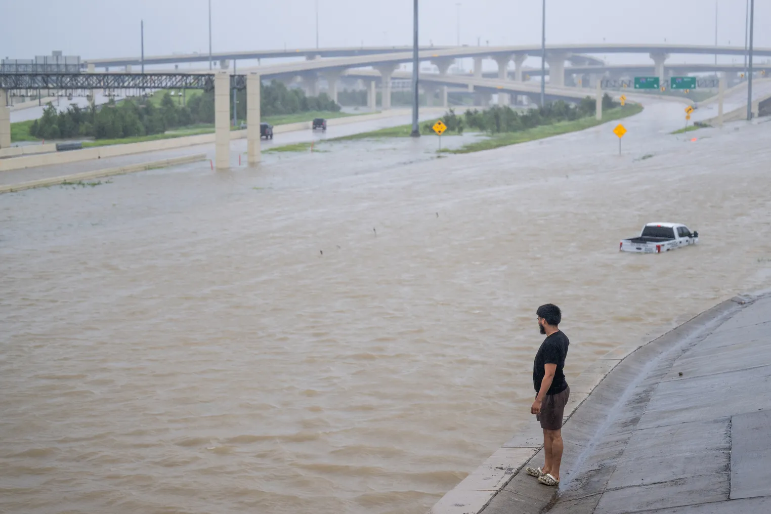 A person looks at a flooded highway with a partly submerged white pickup truck.