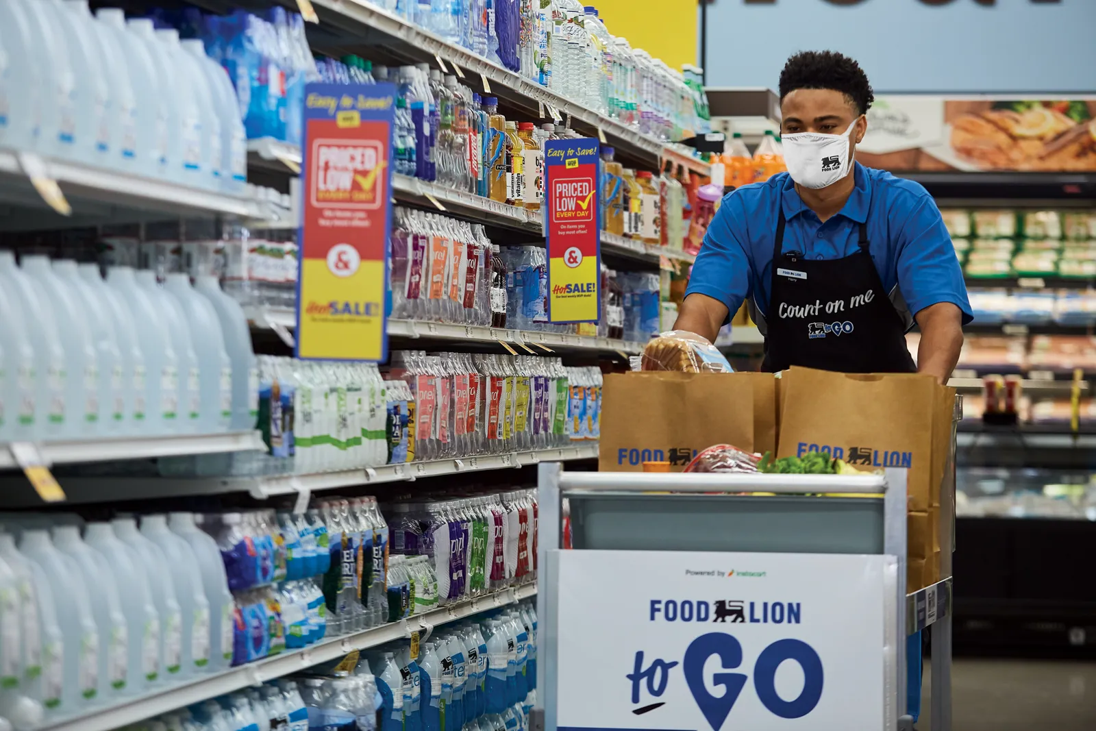 A grocery worker in a blue shirt and black apron that says "Count me" pushes a cart down filled with paper bags and food down a grocery aisle.