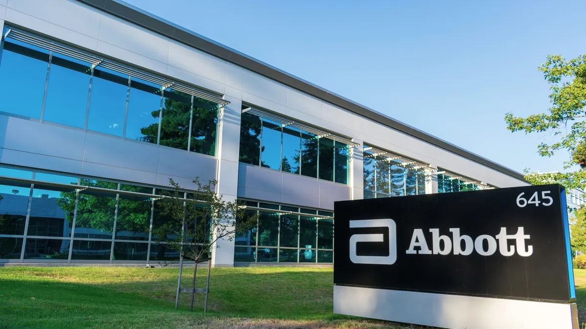 Modern glass and metal office building with a reflective blue-tinted facade, set against a clear blue sky. In the foreground, a large black and white sign reads “Abbott” with the company’s stylized logo and the number “645.” The area has neatly cut grass and a few small trees.