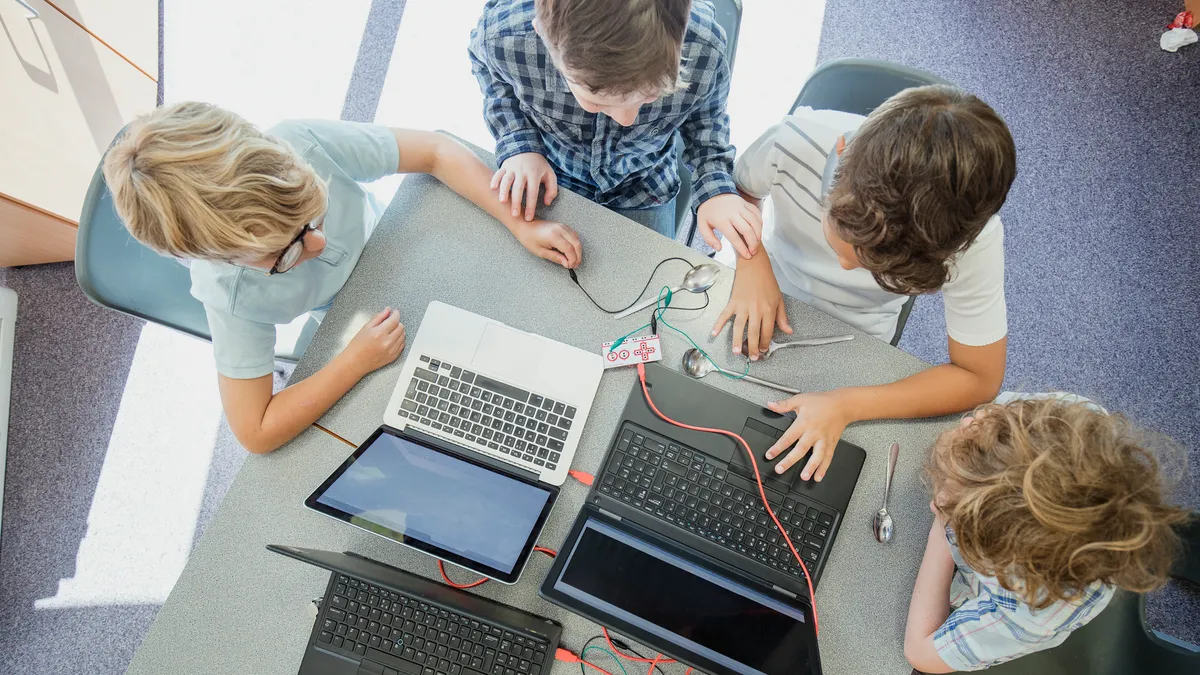 A camera looks down on four students seated at desks, groups together. On the desks are laptops, spoons and wires