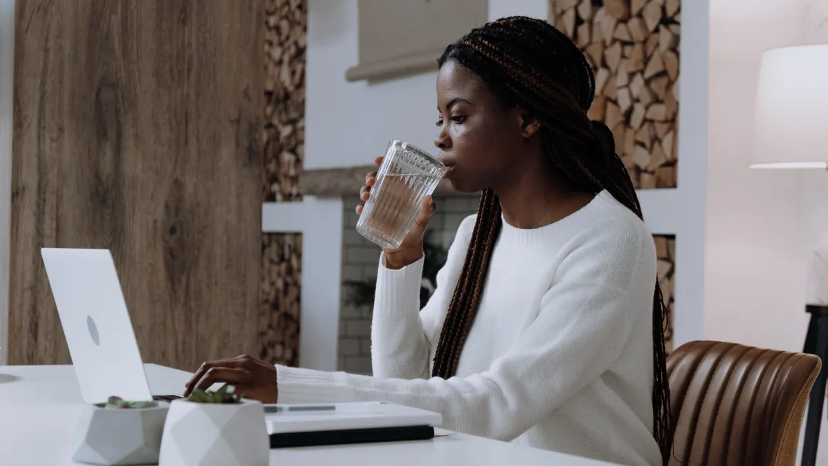 A Black person types on a laptop and sips coffee