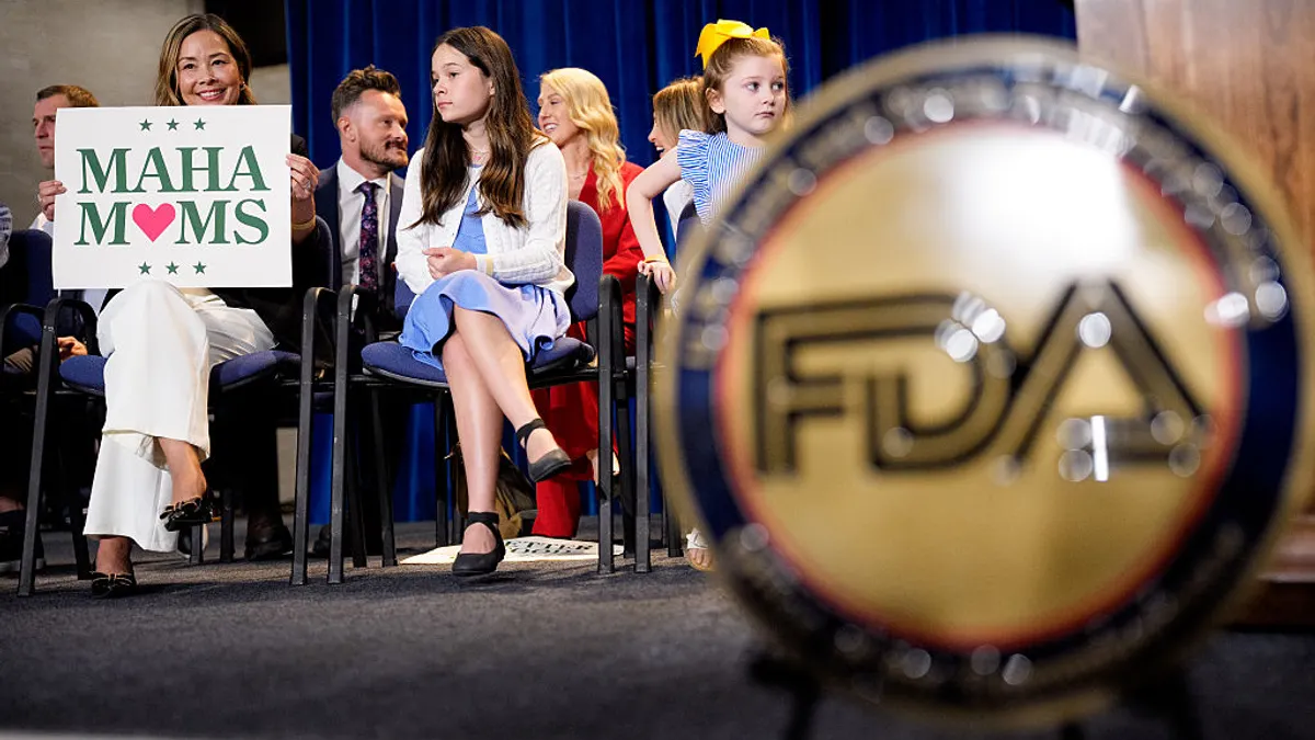 A woman holds a sign that says "MAHA Moms" as she sits on a stage surrounded by children
