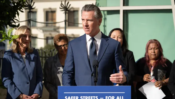 A person in a suit and tie stands behind a microphone at a podium. Behind him, several people watch.