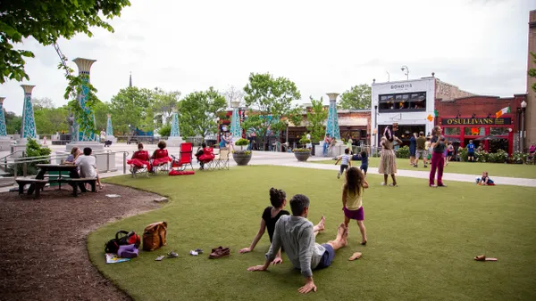 A family of three sits on a patch of green grass as children run and others relax in the distance.