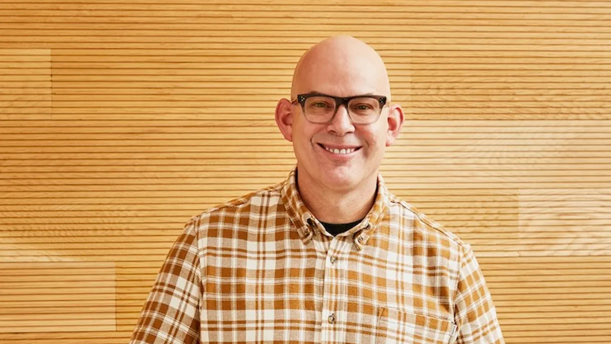 A person with a bald head and eyeglasses wears a yellow and white plaid shirt.