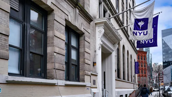 A pedestrian walks down a street with two NYU flags hanging from buildings.