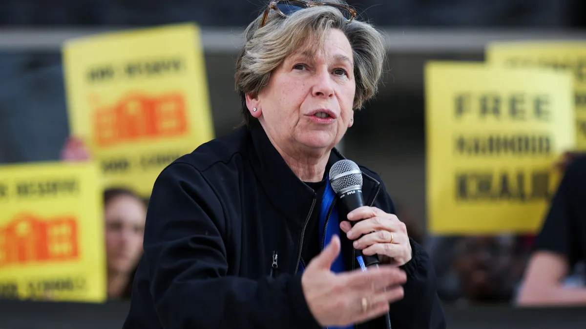 A person speaks using a microphone as they stand in front of a crowd holding yellow protest signs.