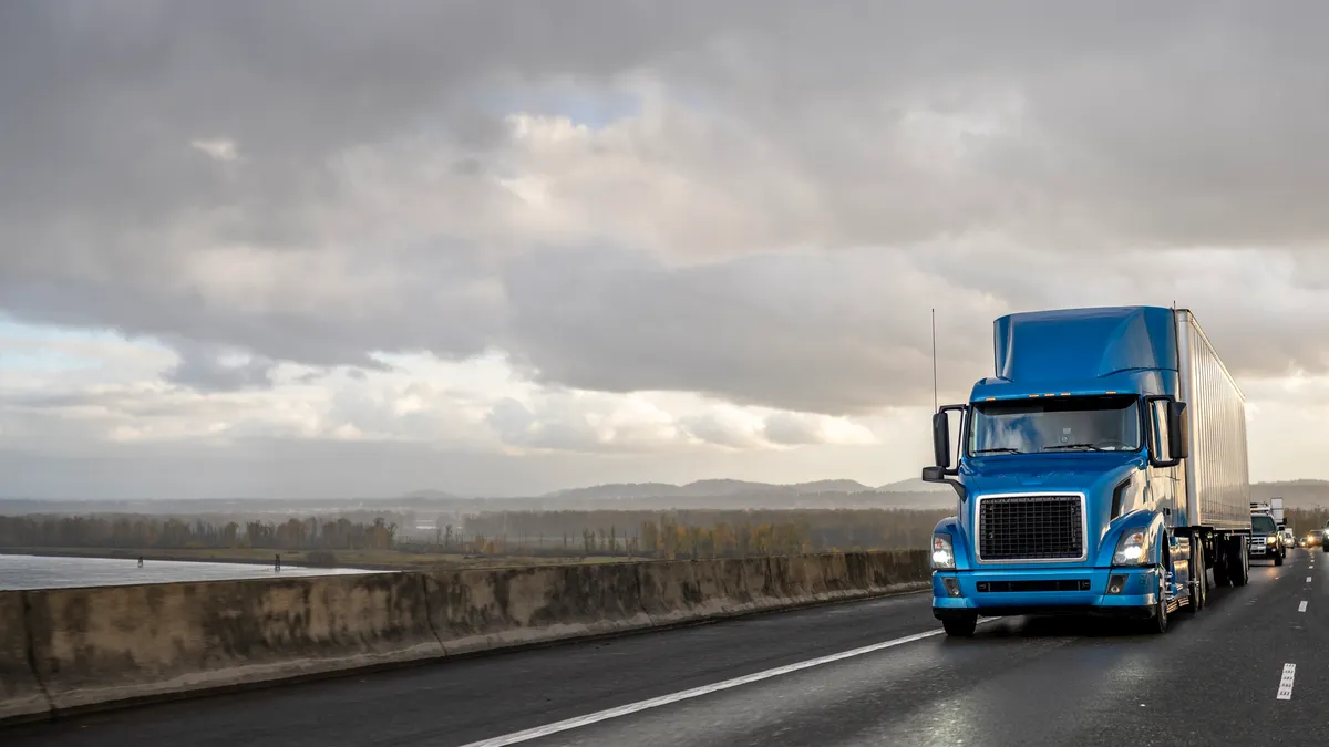 A bright blue tractor-trailer drives on a road under storm clouds.