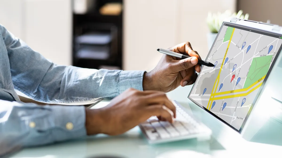 A person's uses a computer keyboard with one hand and points to a map on a screen with the other.