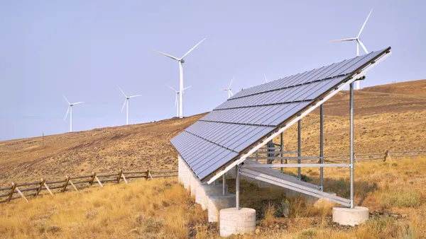 A large solar panel adheres to a cement bass on a hill surrounded by wind turbines. 