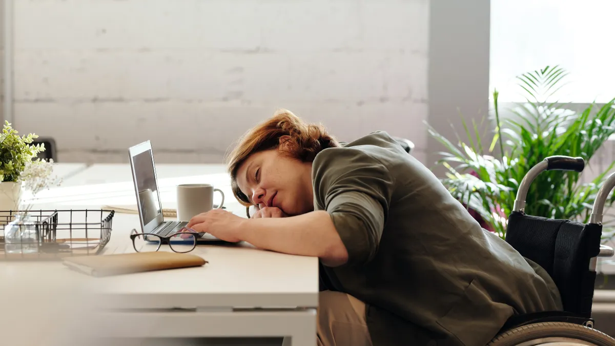 A feminine-presenting person sits in their wheelchair and rests their head on the desk