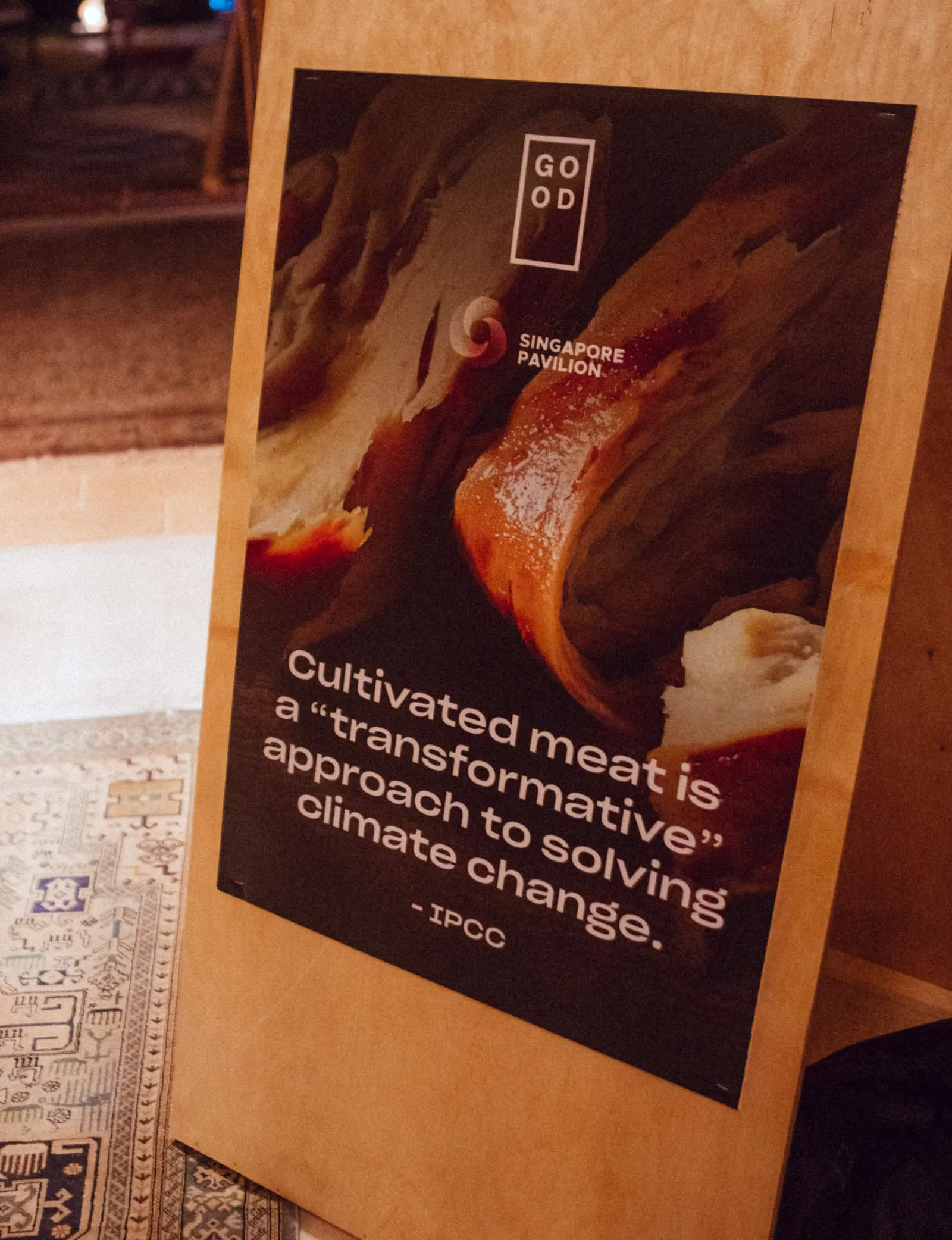 A wooden stand-up sign featuring a quote and image of Good Meat cultivated chicken is on woven rug.