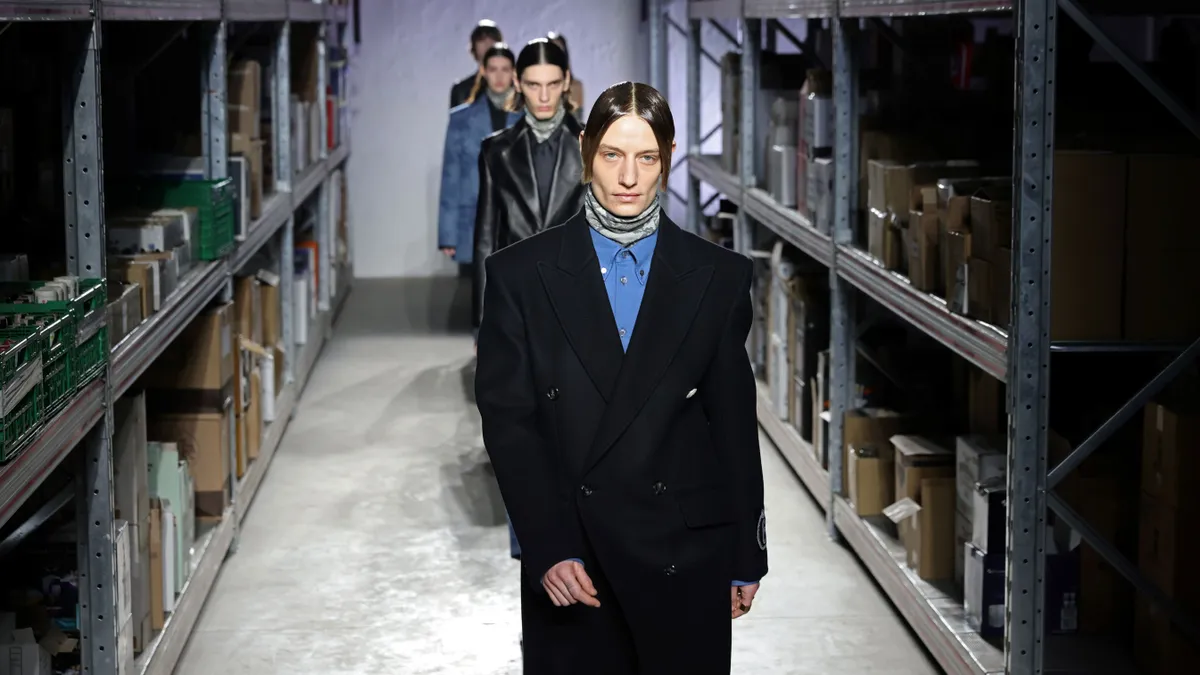 Models walk in a line between metallic warehouse shelves lined with boxes.