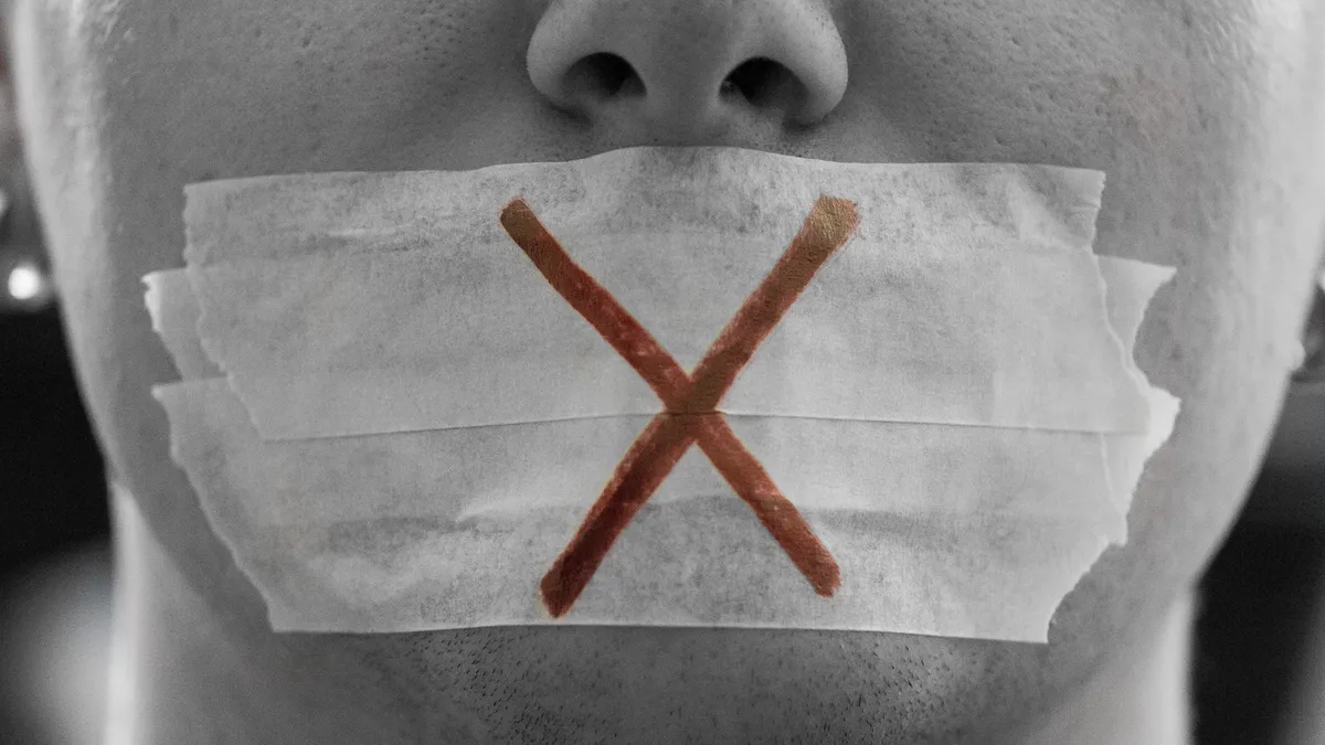 A close up of a person with tape over their mouth and a red "x" across it.