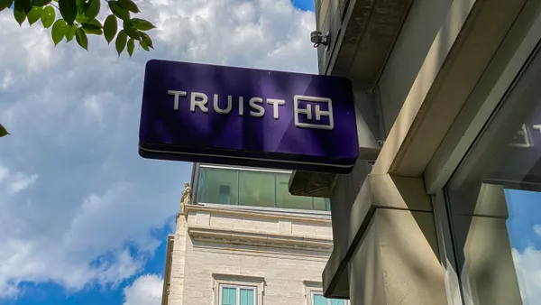 A purple "Truist" bank sign attached to the facade of a stone building is shown, with blue sky and a tall building behind it.