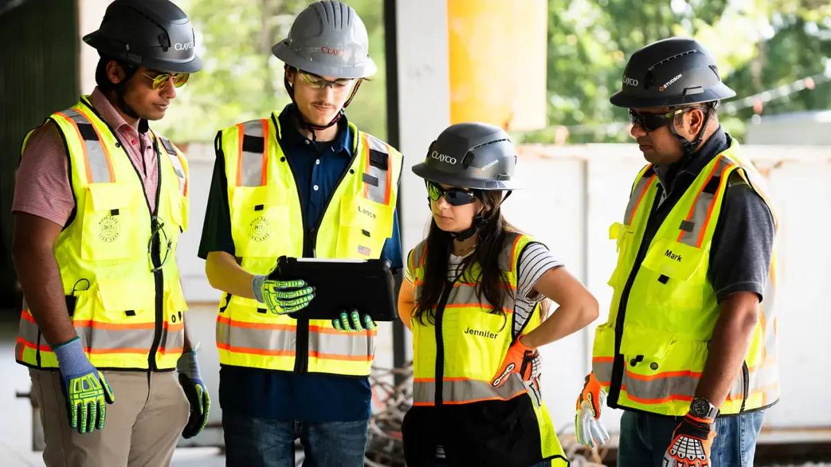 A group of four construction workers stand in a circle and look at a tablet.
