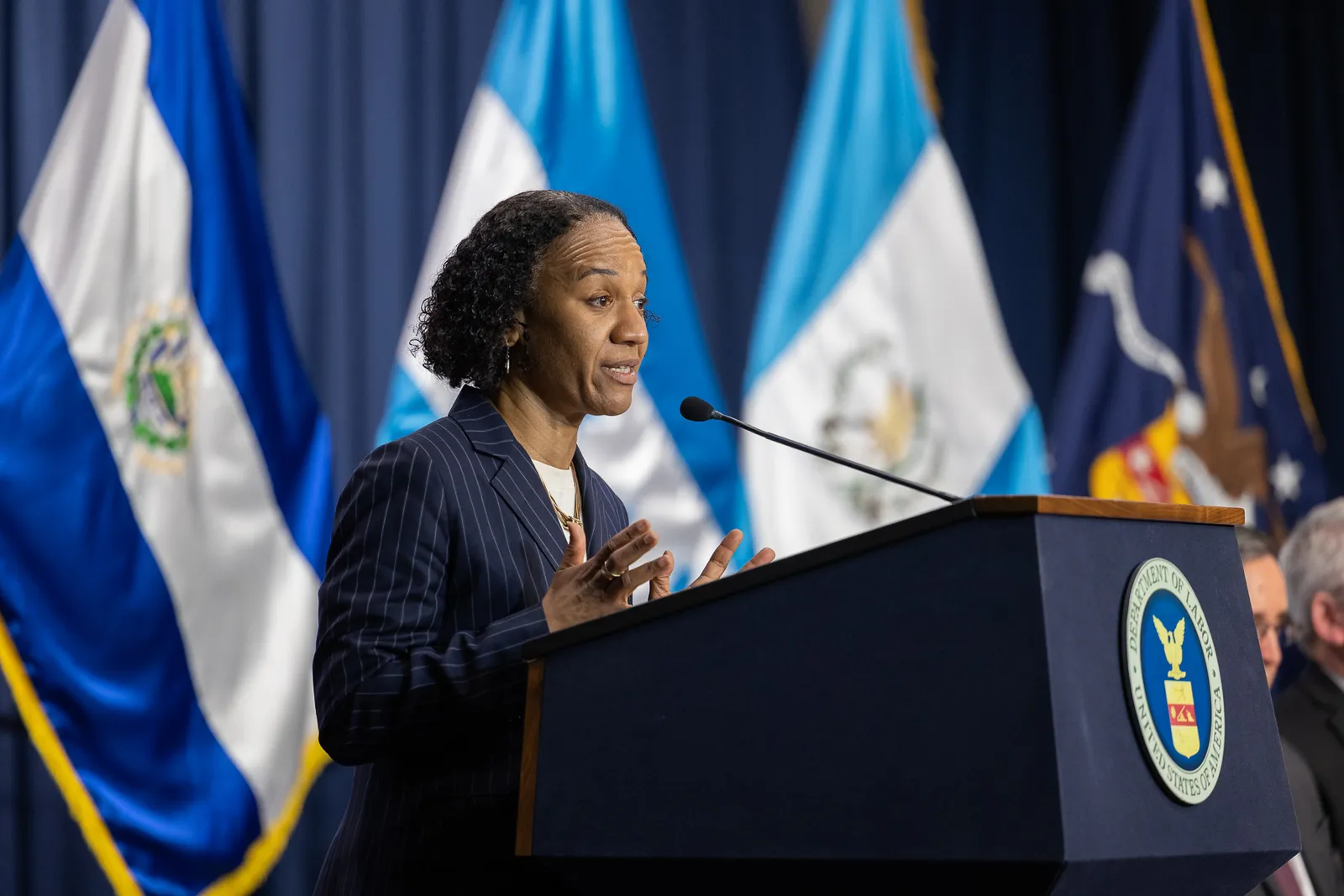 A woman in a suit stands at a podium with a Department of Labor seal on it, speaking.