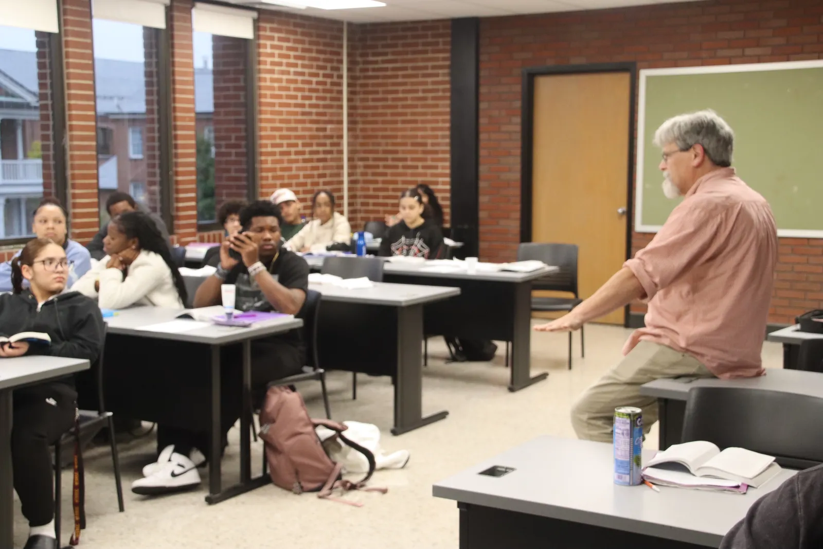 An adult is leaning against a desk in a classroom. Across the aisle from the adult, students are sitting at desks. Some have their heads turned toward the adult.