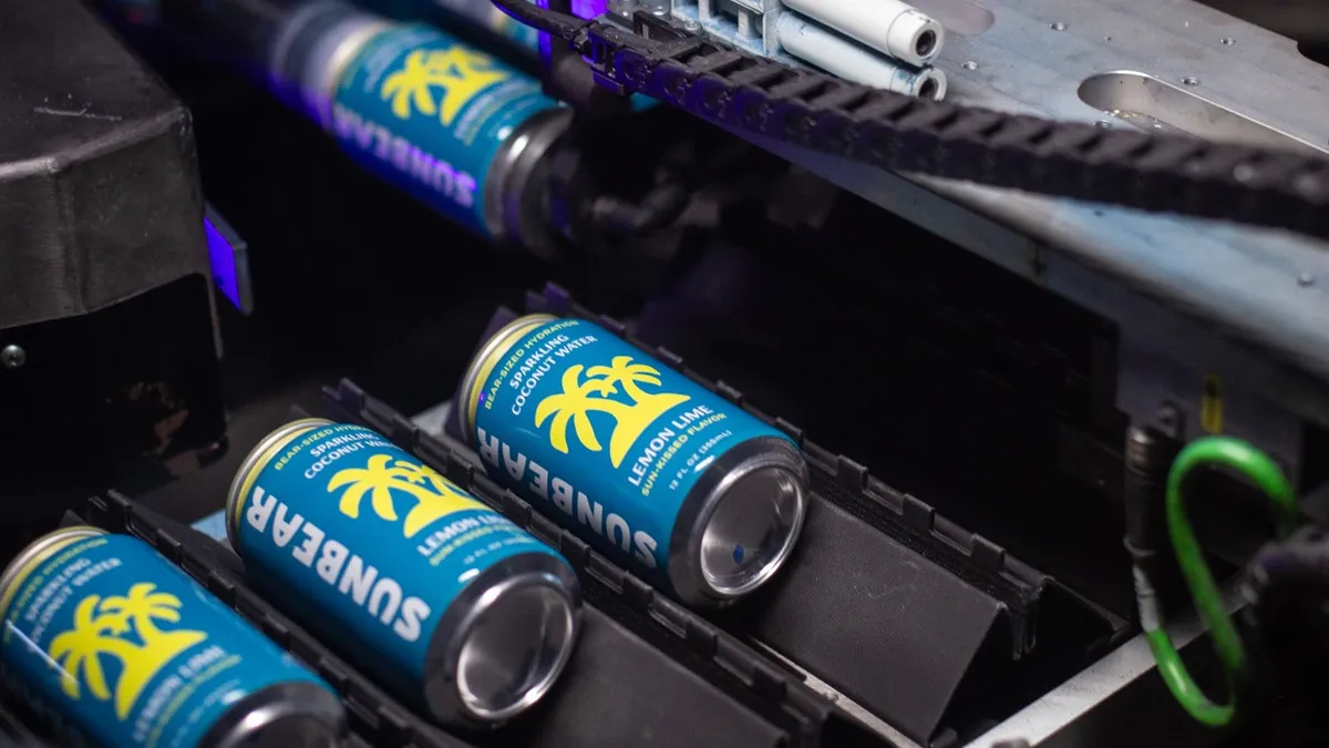 Four 12 fluid-ounce beverage cans laying on a machine with blue and yellow labels that read 'Bear-Sized Hydration; Sparkling Coconut Water; Lemon Lime Sun-Kissed Flavor'