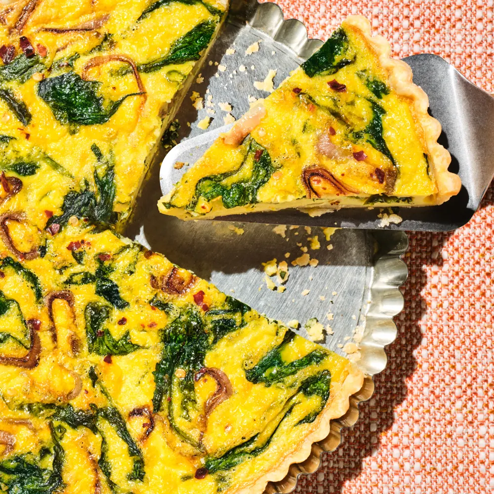 A silver pie server takes a piece of a spinach and vegetable quiche out of a silver pie pan, which is sitting on a woven orange surface.