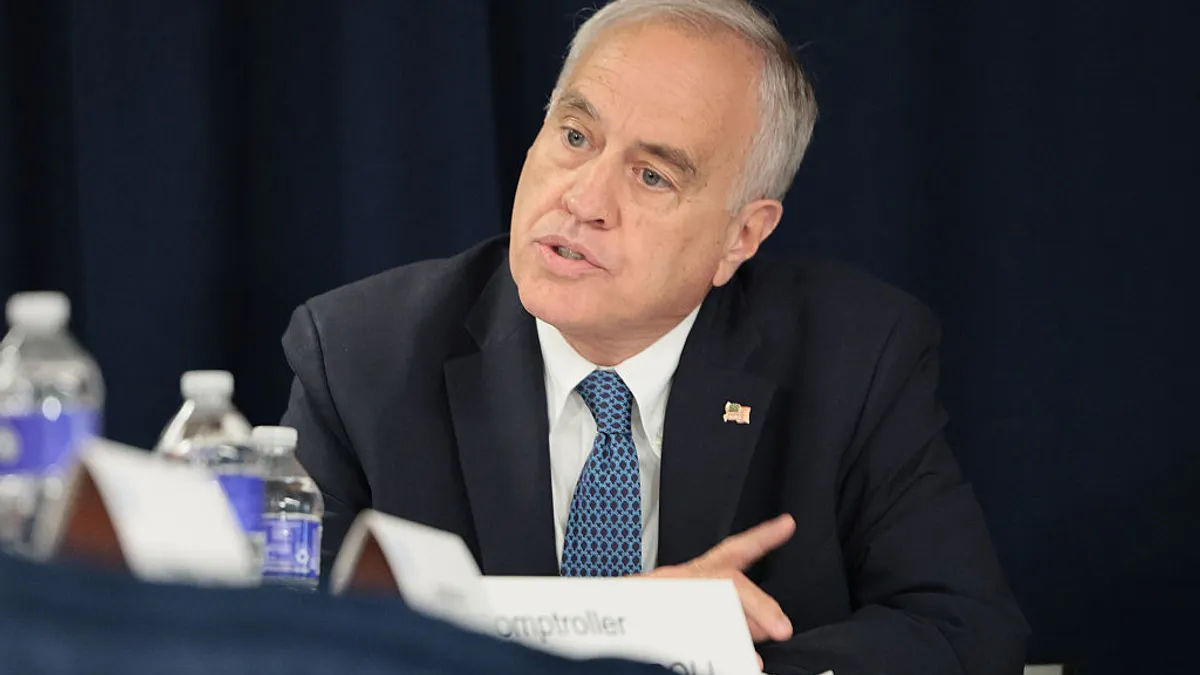 A seated person in a dark suit with a blue tie speaks at a conference table.