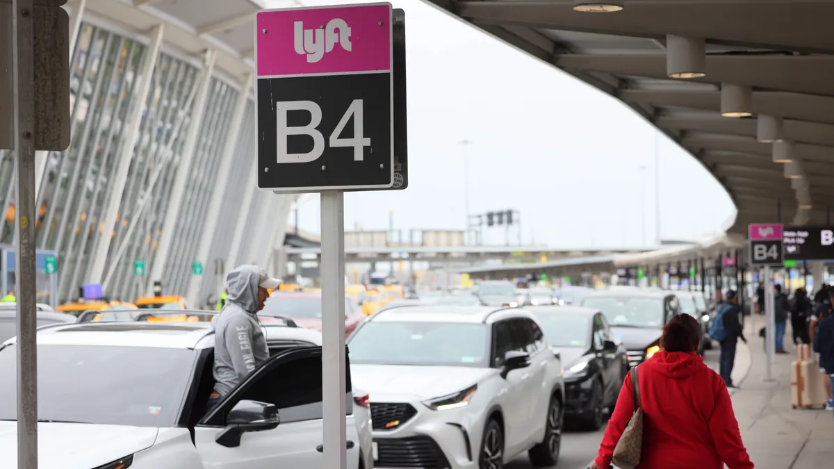 A line of cars under a Lyft ride-hailing sign along a walkway with people and luggage.