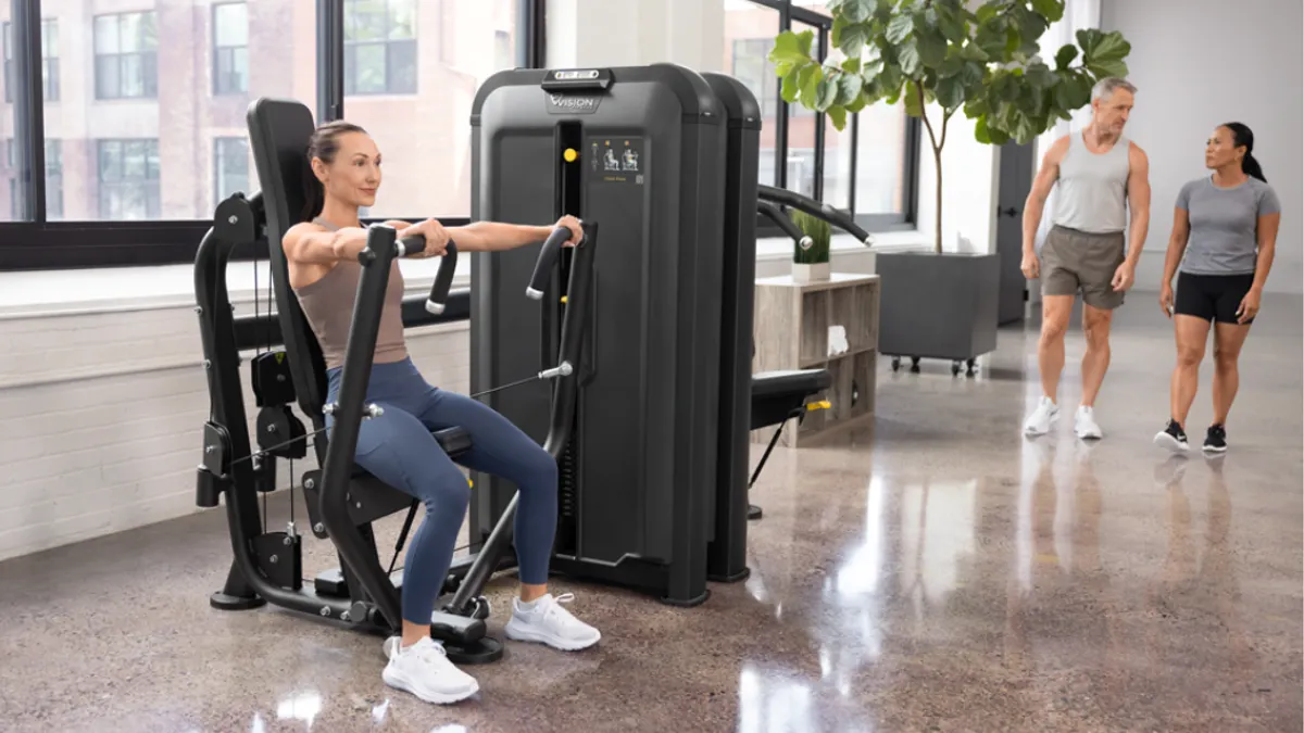 Three people in a gym, with one using a chest press machine while two walk in the background.