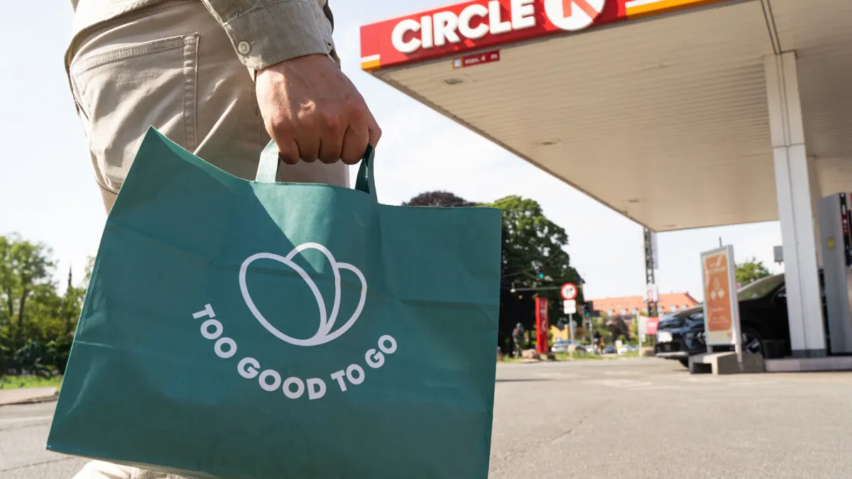 A photo of the lower half of a person, holding a bag and walking toward a gas station. The bag says "Too Good To Go" and the fuel canopy says "Circle K."