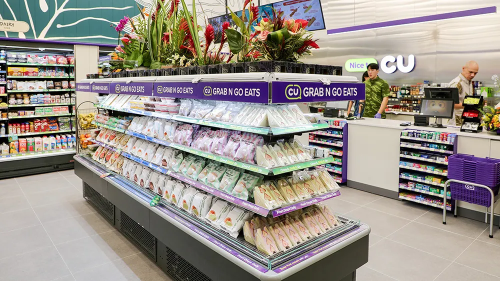 A display of standwiches is arrayed in a cooler in the center of an open space inside a store. The shore is mostly white with accents of purple and teal, and the sandwiches are a variety of fruity and savory options. A riot of tropical flowers adorns the top of the cooler and signs around the top of the cooler say "CU Grab and Go Eats."