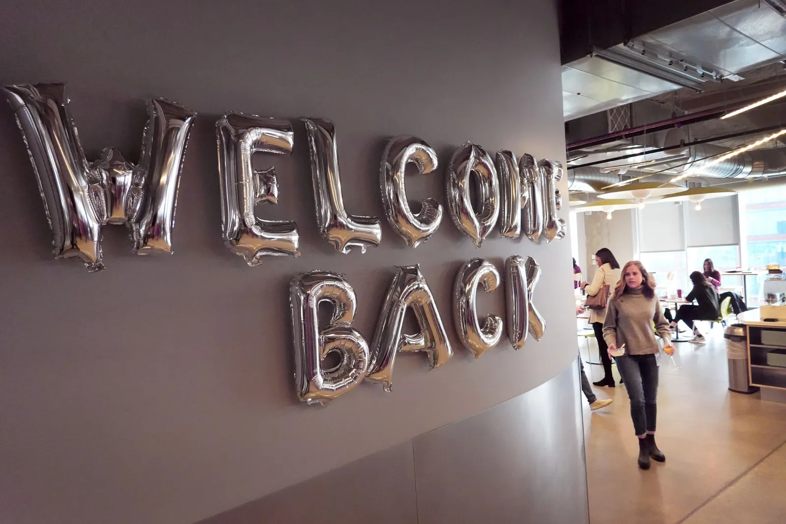 People walk through a Google office cafeteria, past a sign welcoming them back.