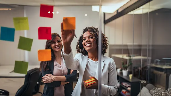 Two women in an office smiling while holding colorful sticky notes, engaged in a collaborative brainstorming session.