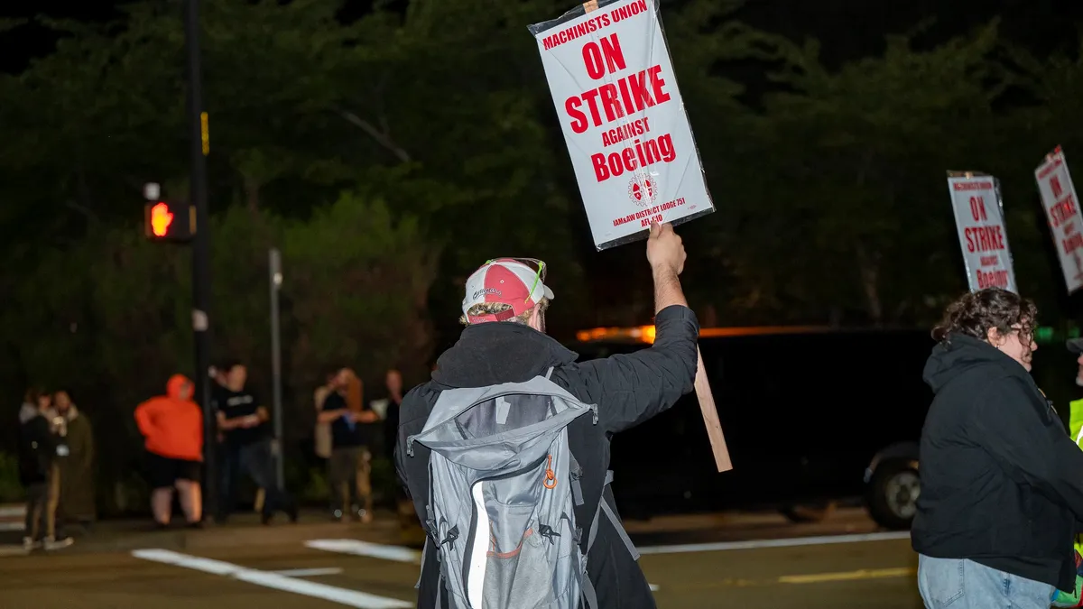 A person's back facing the camera while wearing a black hoodie, gray backpack and a red and white cap holding a red and white strike sign.