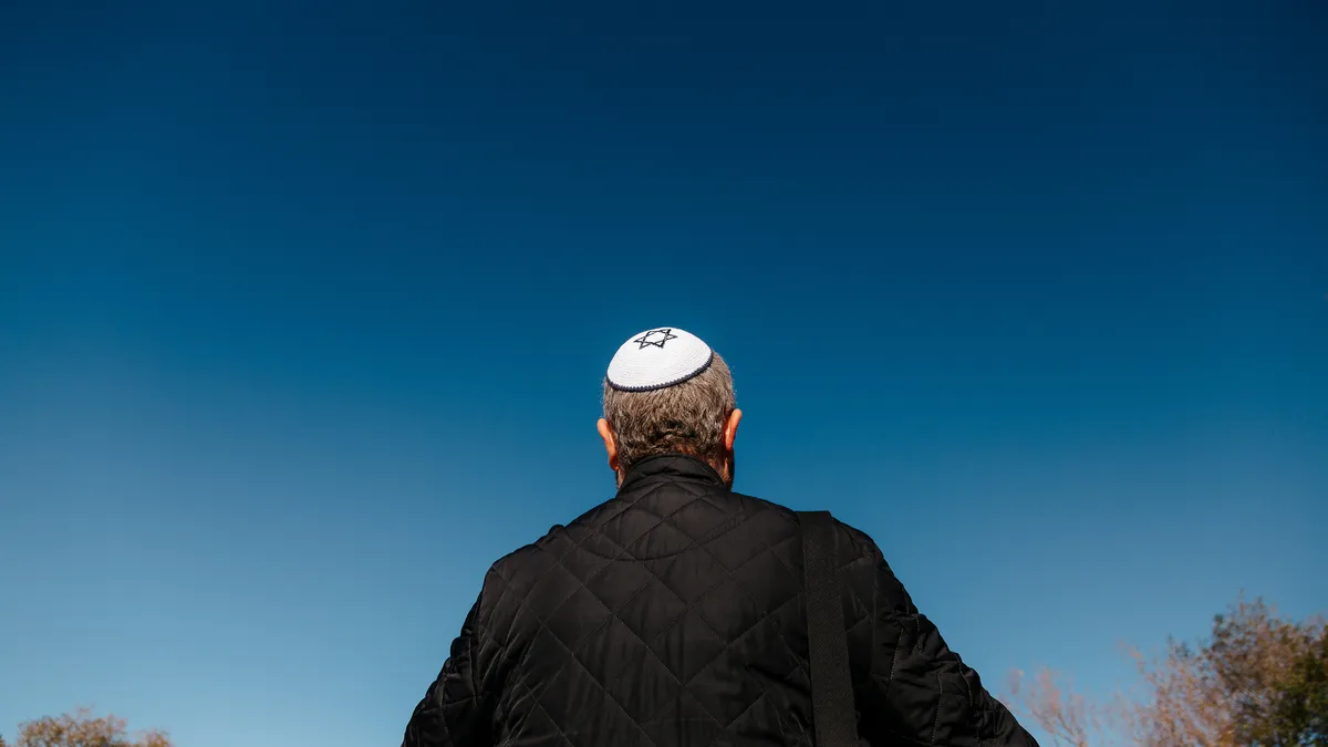 Color image depicting a mid adult Jewish man in his 30s wearing a traditional Jewish skull cap (with star of David design). Rear view low angle image depicting the man looking up at a clear blue sky.