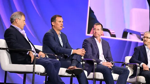 a man seated on a chair flanked by two other men speaks on a keynote stage